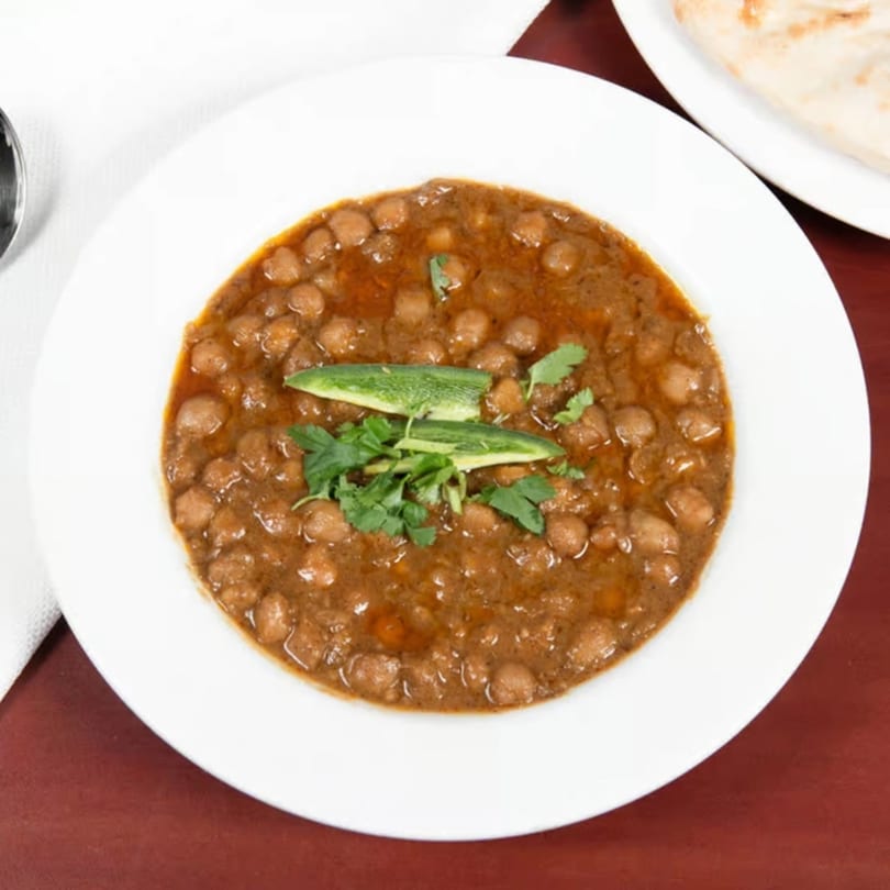 A white bowl of Pakistani chana masala, garnished with green chilies and cilantro, on a red table.